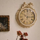 Decorative wall clock with cherub figures on a wall with a framed picture and flowers, Obroi