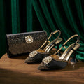 A pair of black pointed-toe slingback high heels with gold studs and crystal embellishments, displayed on a wooden table alongside a matching black studded clutch bag against a dark green velvet backdrop, Obroi