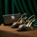 A pair of black pointed-toe slingback high heels with gold studs and crystal embellishments, displayed on a wooden table alongside a matching black studded clutch bag against a dark green velvet backdrop, Obroi