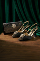 A pair of black pointed-toe slingback high heels with gold studs and crystal embellishments, displayed on a wooden table alongside a matching black studded clutch bag against a dark green velvet backdrop, Obroi