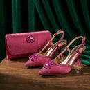 A matching pink formalwear set featuring pointed-toe slingback high heels with rhinestone embellishments and a matching clutch bag, displayed on a wooden surface in front of a dark green velvet curtain, Obroi