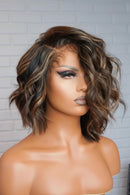 Short wavy wig with highlighted brown and blonde tones, styled on a mannequin head against a light gray brick wall background, Obroi