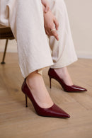A person wearing glossy deep burgundy pointed-toe high heels with slim stiletto heels, paired with cream wide-leg trousers, sitting on a wooden floor, Obroi