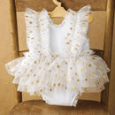 White baby romper with gold star print and ruffled tulle shoulder straps, displayed on a wooden hanger over a wooden chair, Obroi