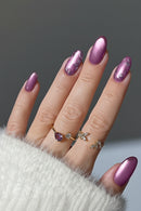 A close-up of a hand with long, glossy mauve-purple manicured nails, wearing two delicate gold rings with small clear stones and a purple heart-shaped stone. The hand is resting on a white fuzzy sleeve against a dark gray background, Obroi