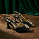 A pair of black pointed-toe slingback heels with gold-tone hardware, rhinestone embellishments, and a sculptural high heel, displayed on a wooden surface with a dark green velvet curtain backdrop, Obroi