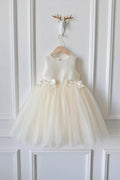 An ivory sleeveless flower girl dress with a tulle skirt and bow details, hanging on a wooden hanger against a white paneled wall, Obroi