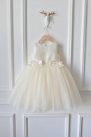 An ivory sleeveless flower girl dress with a tulle skirt and bow details, hanging on a wooden hanger against a white paneled wall, Obroi