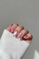 A close-up of a hand with manicured nails decorated in a pastel pink and lavender glitter base, featuring small blue and yellow flower motifs, Obroi