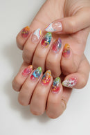 A close-up of a hand showcasing manicured almond-shaped nails with a variety of colorful nail art designs, including floral patterns, rainbow gradients, and small gold star accents, Obroi