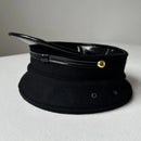 A black military-style peaked cap with a glossy black visor, black leather-like band, and a small gold metal rivet, photographed on a light gray surface, Obroi