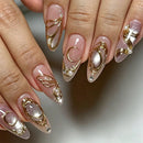 Close-up of manicured fingernails with a glossy nude-pink base, gold metallic swirl and crescent designs, pearl accents, and glitter details, Obroi