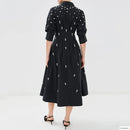 A woman viewed from the back wearing a black midi shirt dress with puff sleeves and scattered white embroidered floral motifs, paired with black heeled sandals against a light gray studio background, Obroi