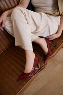 A person seated on a brown leather chair wearing pointed-toe high heels in a deep burgundy patent finish, paired with light beige pants and a beige top, Obroi