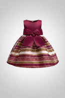 A sleeveless girls' party dress with a voluminous striped skirt and a large satin bow at the waist. The dress features horizontal stripes of deep magenta, glittery gold, and cream, with a solid magenta bodice and bow, Obroi