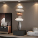 A modern stacked stone sculpture with five smooth rounded stones in white, gray, and peach tones, displayed on a dark gray pedestal in a styled interior setting, Obroi