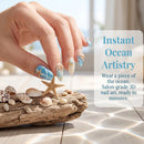 A close-up of a hand with ocean-themed nail art featuring blue starfish and seashell designs, resting near driftwood and shells on a sunlit surface. Text on the right reads 'Instant Ocean Artistry' with promotional copy about ocean-inspired nail art, Obroi