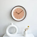 A minimalist round wall clock with a warm peach face, white outer ring, and dark brown frame, mounted on a white wall above a white surface with decorative vases, Obroi