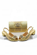 A gold metallic women's formal shoe set with matching clutch handbag, featuring rhinestone-embellished floral details and block heels, displayed on white pedestals against a white background with the brand name Obroi in the top left corner, Obroi