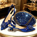 A pair of royal blue satin slingback high-heel pumps with jeweled floral embellishments on the toes, displayed alongside a matching blue and gold beaded clutch purse on a white marble table with gold trim, Obroi
