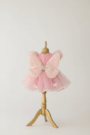 A pink children's dress displayed on a gold dress form, featuring a large butterfly back applique, sheer tulle skirt, and small white butterfly motifs, set against an elegant indoor background, Obroi