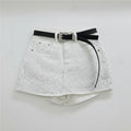 White denim shorts covered in small rhinestones, featuring a black belt with a silver rhinestone-embellished buckle, hanging on a clear plastic hanger against a white wall, Obroi