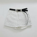 White denim shorts covered in small rhinestones, featuring a black belt with a silver rhinestone-embellished buckle, hanging on a clear plastic hanger against a white wall, Obroi
