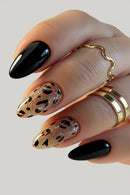 A close-up of a hand with manicured nails, featuring two glossy black nails and two glittery nude-brown nails with black leopard spots. The hand is adorned with gold rings against a light gray background, Obroi