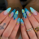 Close-up of hands with long, sculpted almond-shaped nails featuring blue and turquoise floral designs, sheer pink-to-clear gradient tips, and small gold star embellishments. Rings are worn on the fingers, and tattoos are visible on the left hand, Obroi