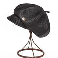 Black quilted faux-leather newsboy cap with a small gold-tone button detail, displayed on a brown wire stand against a white background, Obroi