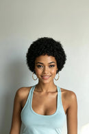 A smiling woman with short, curly black hair wearing a light blue tank top and gold hoop earrings, photographed against a plain light gray wall, Obroi