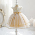 A toddler girl's formal party dress displayed on a white mannequin. The dress features a cream lace bodice with gold floral embroidery, short puff sleeves, a matching cream tulle skirt, a central gold ribbon bow at the waist, and a layered tulle skirt with gold trim, Obroi