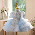 A light blue and white baby girl's tutu dress displayed on a gold dress form mannequin. The dress features a light blue tulle skirt, a white bodice with intricate silver beadwork and embroidery, and short puff sleeves, Obroi