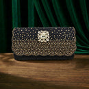 A black evening clutch bag heavily embellished with gold rhinestones and a large central crystal-encrusted clasp, displayed on a wooden surface in front of a dark green velvet curtain, Obroi