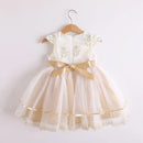 A toddler girl's formal dress with a cream lace bodice, gold embroidered details, and a tiered tulle skirt in champagne beige with a large gold ribbon bow at the waist, hanging on a wooden hanger against a plain light background, Obroi