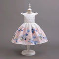 A white girl's party dress displayed on a white dress form mannequin against a solid gray studio background. The dress features a white bodice with tiny silver star embellishments, a sheer lace waistband, and a full flared skirt printed with a watercolor-style floral pattern in shades of blue, pink, and green, Obroi