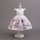 A white girl's party dress displayed on a white dress form mannequin against a solid gray studio background. The dress features a white bodice with tiny silver star embellishments, a sheer lace waistband, and a full flared skirt printed with a watercolor-style floral pattern in shades of blue, pink, and green, Obroi
