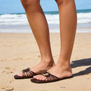 Person wearing sandals on a sandy beach with ocean waves in the background, Obroi