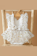 White sleeveless baby dress with gold star pattern, ruffled shoulders, a bow at the chest, and a tulle tutu skirt, displayed on a wooden hanger against a rustic wooden chair, Obroi