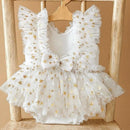 White sleeveless baby dress with gold star pattern, ruffled shoulders, a bow at the chest, and a tulle tutu skirt, displayed on a wooden hanger against a rustic wooden chair, Obroi