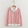 A pink long-sleeve V-neck sweatshirt with a white pointed collar and a small embroidered cat motif on the chest, hanging on a wooden hanger against a light wall, Obroi