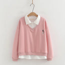 A pink long-sleeve V-neck sweatshirt with a white pointed collar and a small embroidered cat motif on the chest, hanging on a wooden hanger against a light wall, Obroi