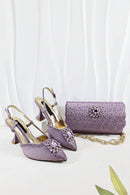 A matching purple women's accessory set featuring a pair of pointed-toe slingback high heels and a textured clutch bag with rhinestone embellishments and a gold chain strap, displayed on a white draped fabric background, Obroi