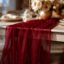 Red sheer fabric with beading on a table with blurred floral arrangement in the background, Obroi