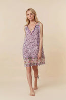 A woman modeling a sleeveless V-neck mini dress with a purple and pink floral pattern and a decorative border, set against a solid beige background, Obroi