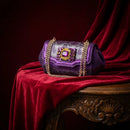 A purple textured handbag with a gold chain strap and a jeweled rectangular clasp, displayed on a draped red velvet surface over an ornate wooden table, Obroi
