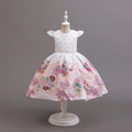 A white girls' party dress displayed on a white dress form against a solid gray studio background. The dress features a white bodice with small silver rhinestone embellishments, a thin silver waistband, and a full pink floral skirt with purple, red, and green flowers, Obroi