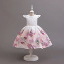 A white girls' party dress displayed on a white dress form against a solid gray studio background. The dress features a white bodice with small silver rhinestone embellishments, a thin silver waistband, and a full pink floral skirt with purple, red, and green flowers, Obroi