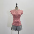A pink short-sleeve top with black polka dots is displayed on a mannequin, with five additional color options (burgundy, beige, white, and black) hanging on a rack behind it against a white louvered screen, Obroi