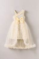 A sleeveless cream-colored baby dress with a textured knit bodice, a large matching bow at the waist, and a layered tulle skirt with ruffled trim, shown against a plain light gray background, Obroi